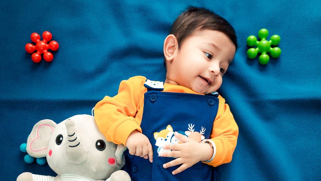 A cute baby boy lying on a vibrant blue blanket with a stuffed elephant toy and colorful toys.