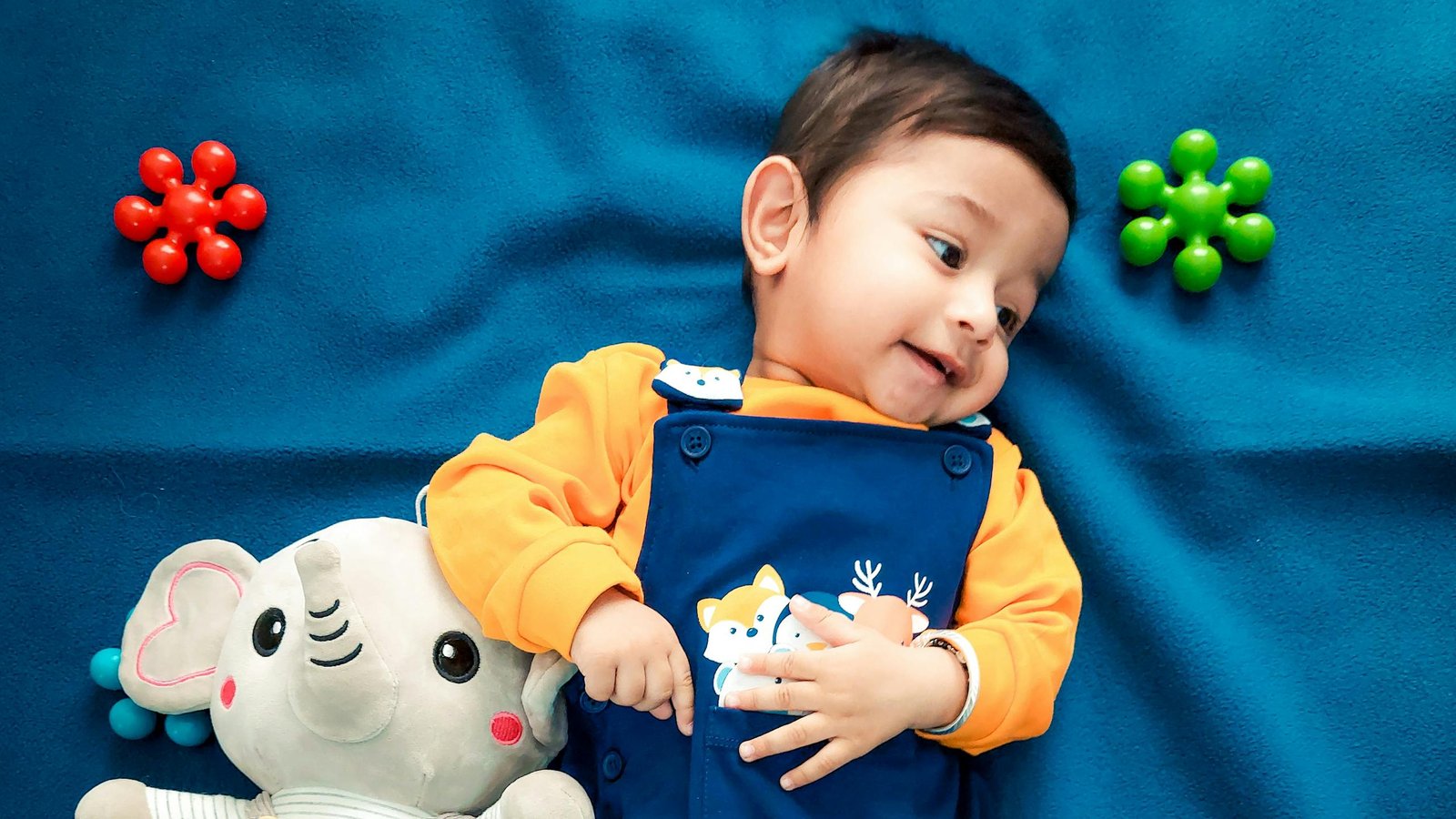 A cute baby boy lying on a vibrant blue blanket with a stuffed elephant toy and colorful toys.