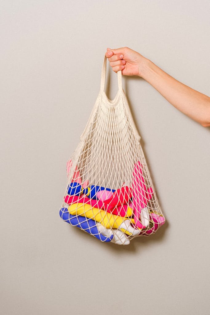 A mesh bag filled with colorful silicone sex toys held by an adult's hand against a neutral background.